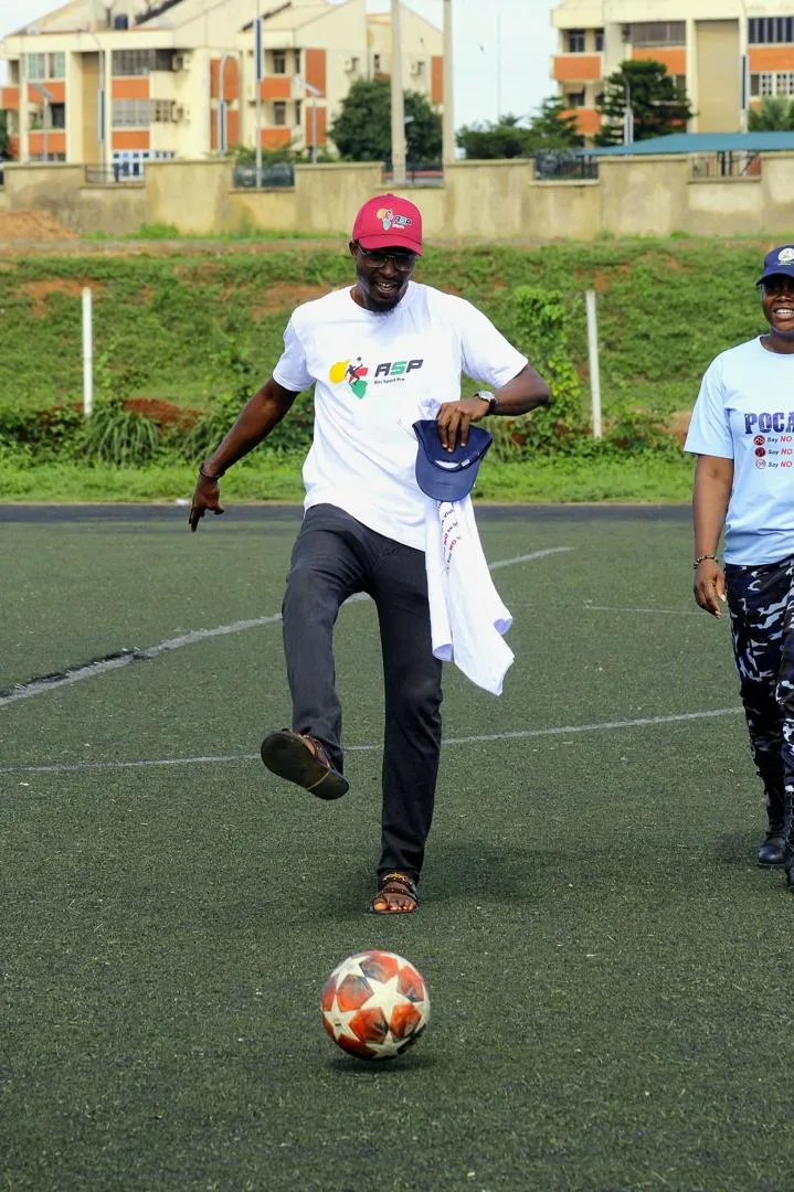 A man in a red cap and an ASP Afri Sport Pro T-shirt kicking a football on an artificial pitch.