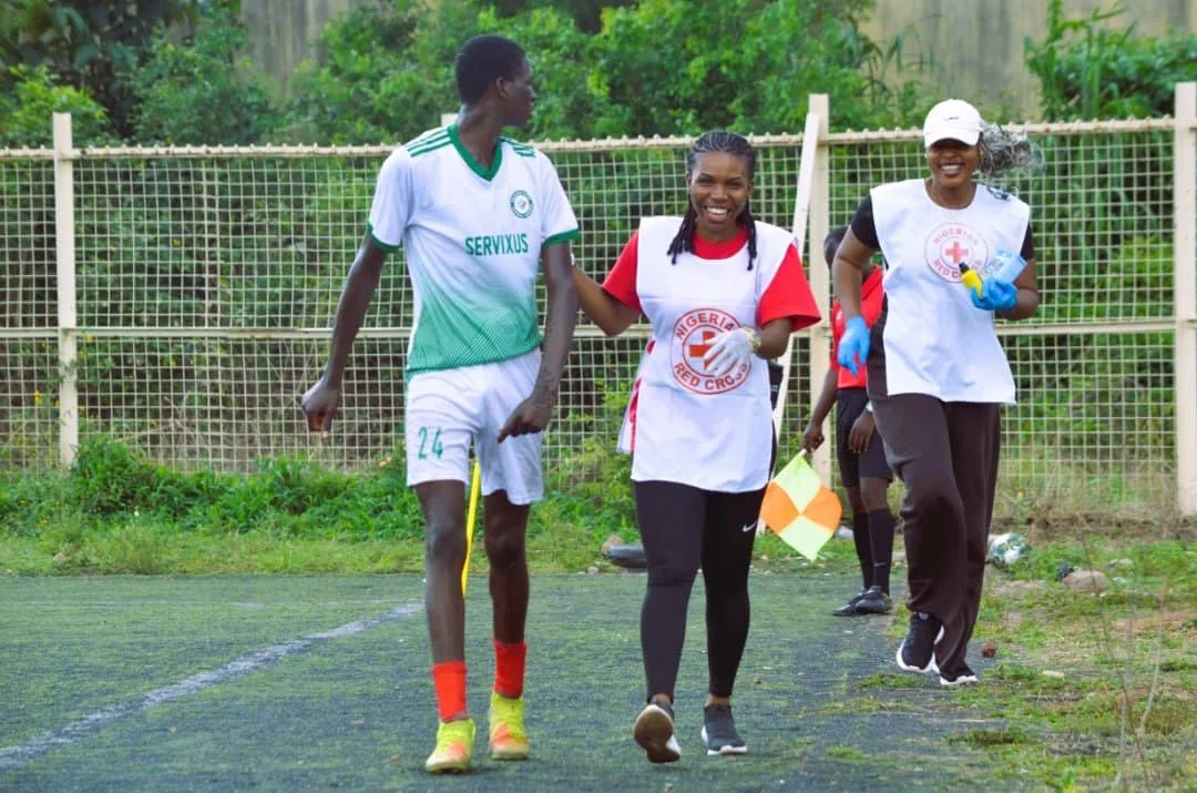 A young player in a green and white kit walking off the pitch beside two smiling Nigerian Red Cross volunteers, an assistant referee’s flag behind them.