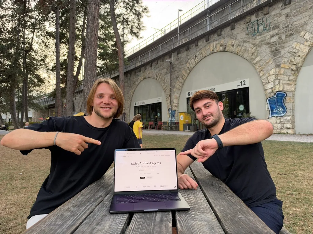 Yves Zumbühl and Claudio Berardi at a wooden picnic table beneath a stone railway viaduct, both pointing at a laptop showing the botts.ai website.