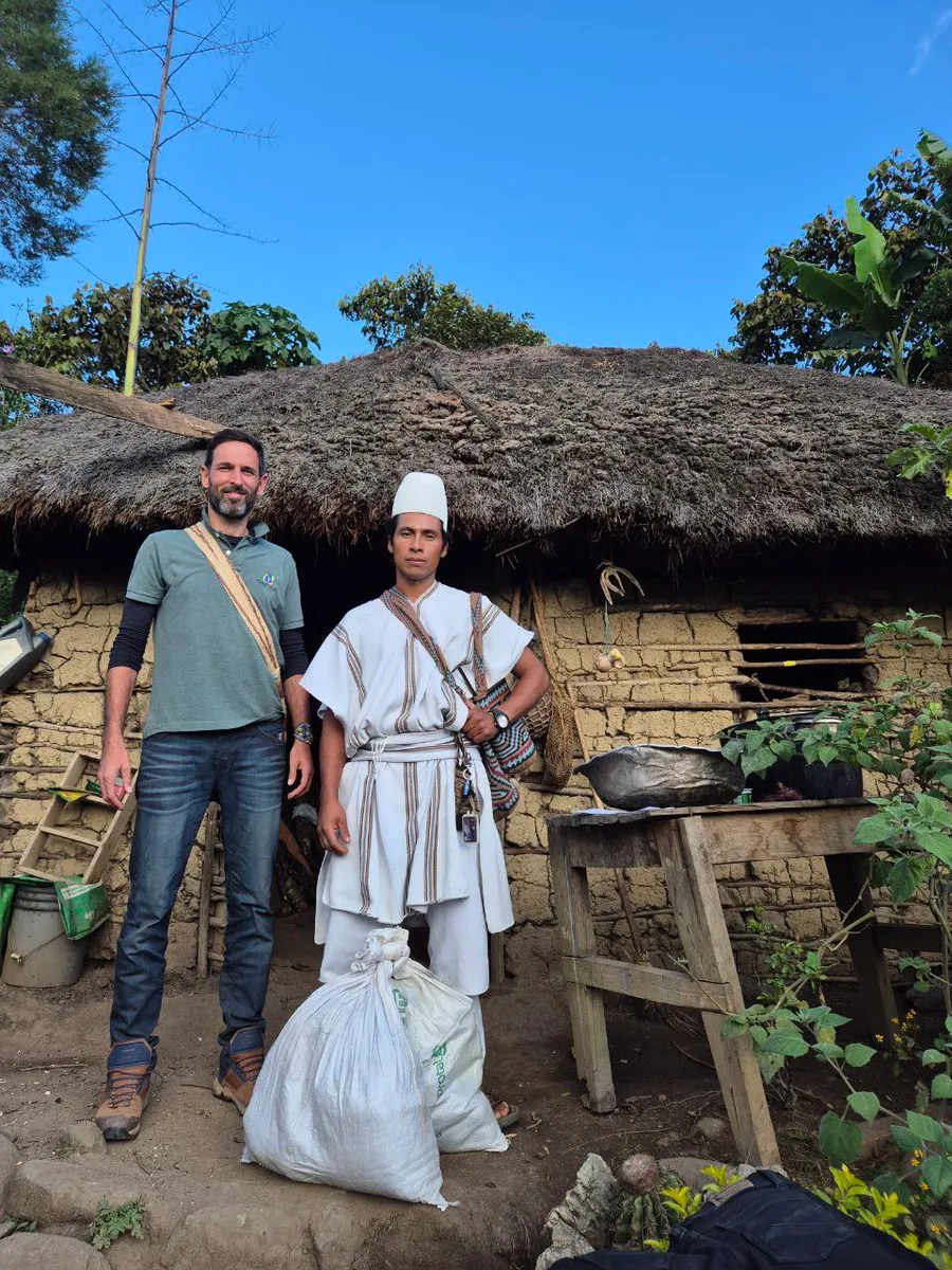 Laurent Gheller standing beside a man in white traditional dress in front of a thatched hut, sacks at their feet.