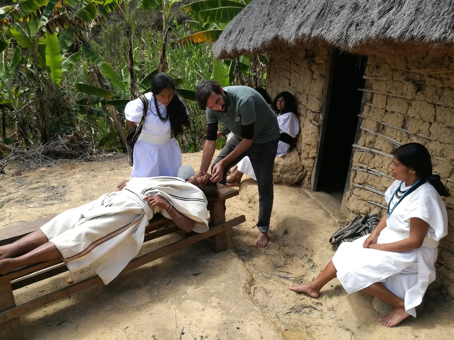 Laurent Gheller leaning over a man lying on a wooden bench outside a thatched mud-walled house, with three women in white traditional dress beside them.
